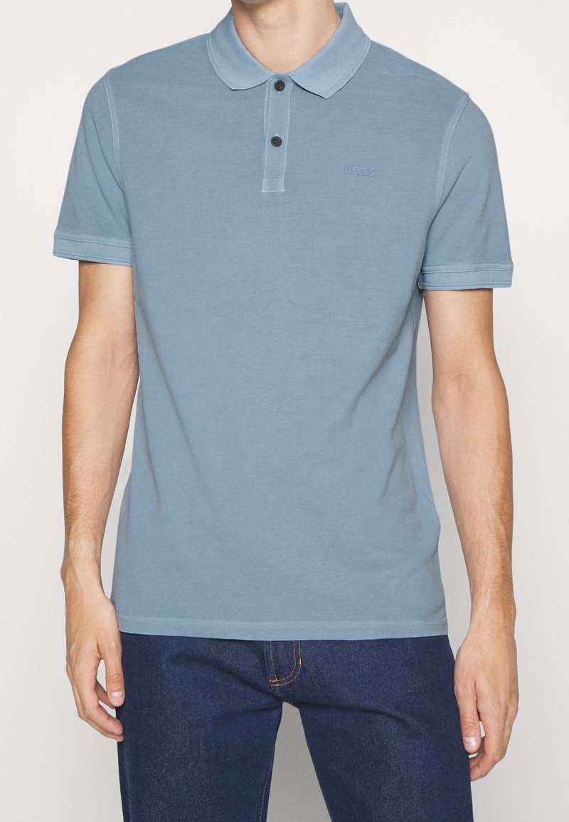Homme portant un polo bleu clair à manches courtes avec deux boutons et un jean bleu foncé, debout les bras détendus.