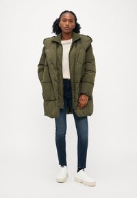 Manteau rembourré vert olive avec un col haut, des boutons sur le devant et une fermeture éclair. Porté sur un pull crème, associé à un jean foncé et des baskets blanches.
