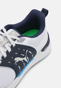 Hvid sportsko med marineblå detaljer, mesh-overdel, tekstureret tunge og grå branding. Har snørebåndsdesign og polstret grøn indersål.