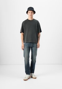 Camiseta oversized gris oscuro, desgastada con cuello redondo; jeans azul claro; zapatillas blancas con suelas marrones; sombrero tipo bucket negro.