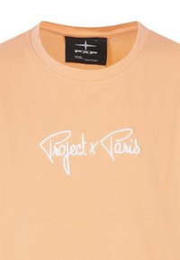 T-shirt en coton orange avec un col rond, arborant le texte brodé en blanc "Project x Paris" à l'avant. Texture lisse, coupe décontractée.