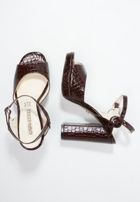 Crocodile-patterned burgundy high-heeled sandals with an open toe, wide strap, and adjustable ankle buckle. Block heel design.