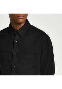 Czarna koszula na guziki wykonana z teksturowanej tkaniny, z dwoma przednimi kieszeniami, szpiczastym kołnierzykiem i czarnymi guzikami.