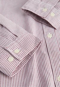 Gros plan d'une chemise à manches longues pliée avec de fines rayures verticales rouges et blanches et des boutons blancs aux poignets et sur la patte de boutonnage avant.
