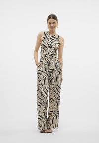 Beige Jumpsuit mit schwarzen Zebra-Streifen, mit ärmellosem Design, lässigem Sitz, elastischem Bund und seitlichen Taschen. Dazu werden schwarze Sandalen getragen.