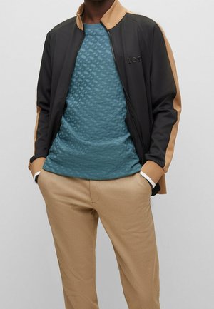 Mann trägt schwarze und braune Zip-up-Jacke über teal texturiertem Hemd mit beigen Hosen, Hände in den Taschen vor einfarbigem Hintergrund.