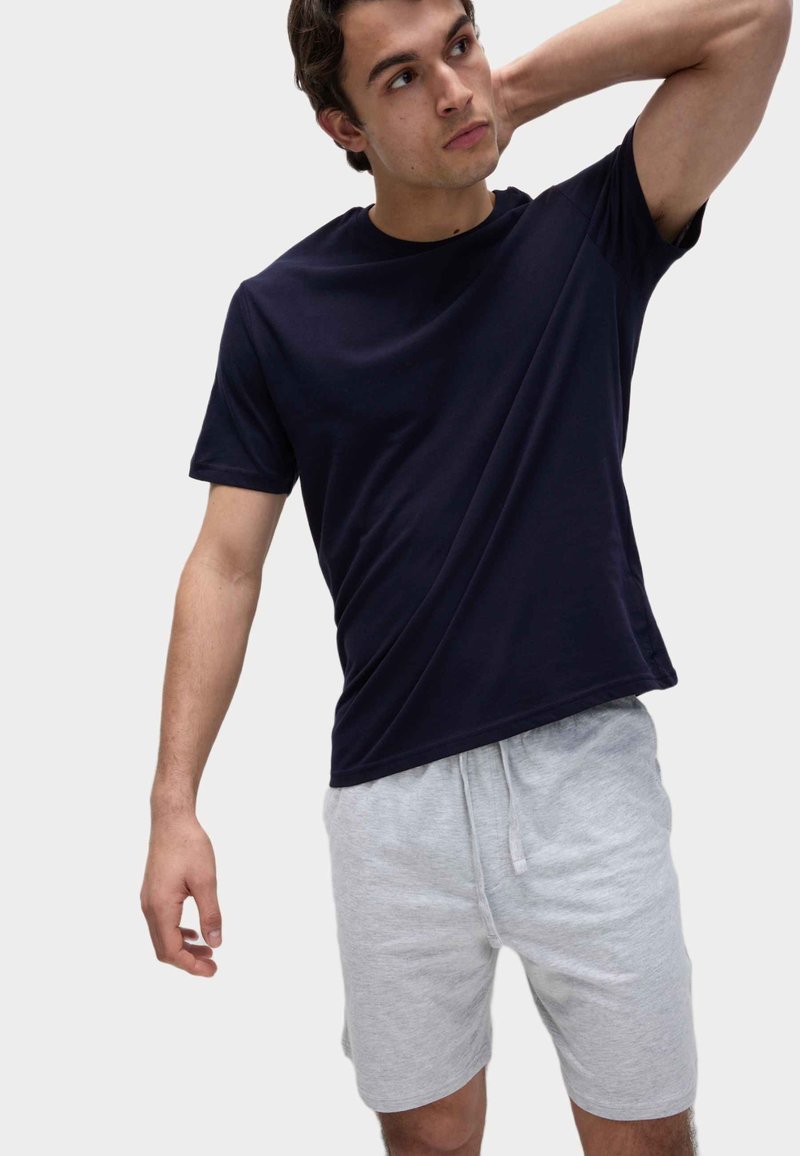 Giovane uomo con maglietta blu navy e pantaloncini grigi chiaro con coulisse, che posa con un braccio sollevato dietro la testa.