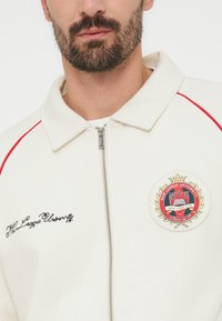 Άνδρας με γένια που φοράει λευκό μπουφάν με φερμουάρ και κόκκινο ρέλι, κεντημένο κείμενο "Νέα Λιγκ Πανεπιστήμιο" και στρογγυλό έμβλημα NLU Νέα Υόρκη.