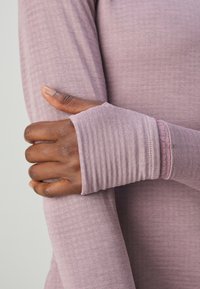 T-shirt à manches longues en tissu doux de couleur violet clair avec texture côtelée. Présente des poignets sans doigts. Bras positionné à travers le corps.