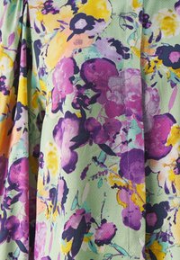 Robe florale avec un fond vert pastel et des fleurs violet, jaune et rose vives. Tissu doux et texturé avec des plis.