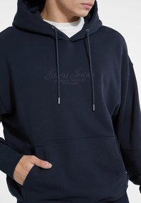 Sudadera con capucha de color azul marino, hecha de tela texturizada, que cuenta con un bolsillo frontal, bordado del logo y cordones ajustables con puntas de metal.