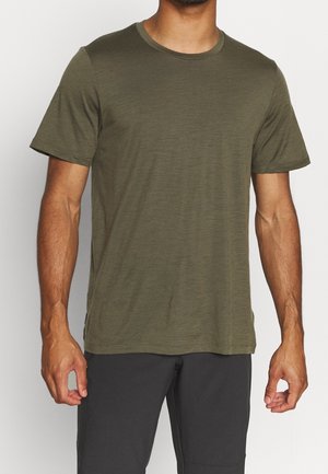 Homme portant un t-shirt olive à manches courtes et col rond, ainsi qu'un pantalon gris foncé, debout avec les bras détendus le long du corps.