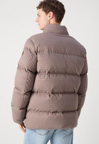 Casaco puffer em padrão xadrez castanho e bege, com um colarinho alto e design volumoso, apresentando costuras visíveis e logotipo na manga.