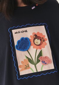 Tumesinine T-särk, millel on ristküliku kujuline graafika koos karu, lillede ja värviliste heegeldatud aplikatsioonidega sinistes ning roosades toonides; ääristatud lainelise paelaäärisega.