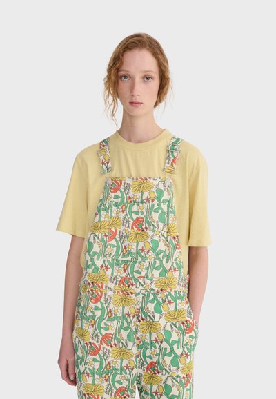 Jeune personne aux cheveux roux bouclés portant une chemise jaune à manches courtes sous une salopette à motifs floraux avec des fleurs vertes, jaunes et rouges.
