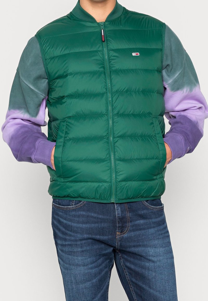 Homme portant un gilet matelassé vert zippé sur un sweat à dégradé violet et vert, les mains dans les poches du gilet et un jean bleu.