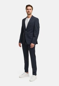 Abito blu navy con giacca su misura e pantaloni slim-fit, abbinato a una maglietta bianca e sneakers bianche. Tessuto morbido e linee pulite.