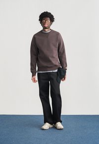 Σκούρο καφέ φούτερ με ριμπ μανσέτες, φορεμένο πάνω από ένα ανοιχτό γκρι πουκάμισο, σε συνδυασμό με μαύρο παντελόνι και κρεμ αθλητικά παπούτσια, με αξεσουάρ μαύρο καπέλο.