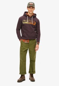 Ciemnofioletowa bluza z kapturem z logo "Superdry", założona na koszulę w kratę. Oliwkowe spodnie i brązowe wiązane buty dopełniają całość.