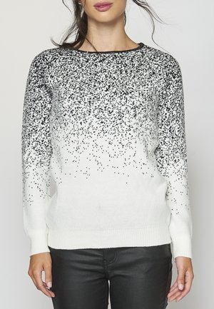 Femme vêtue d'un pull blanc avec un motif tacheté noir qui s'estompe des épaules vers le bas, associé à un pantalon noir.