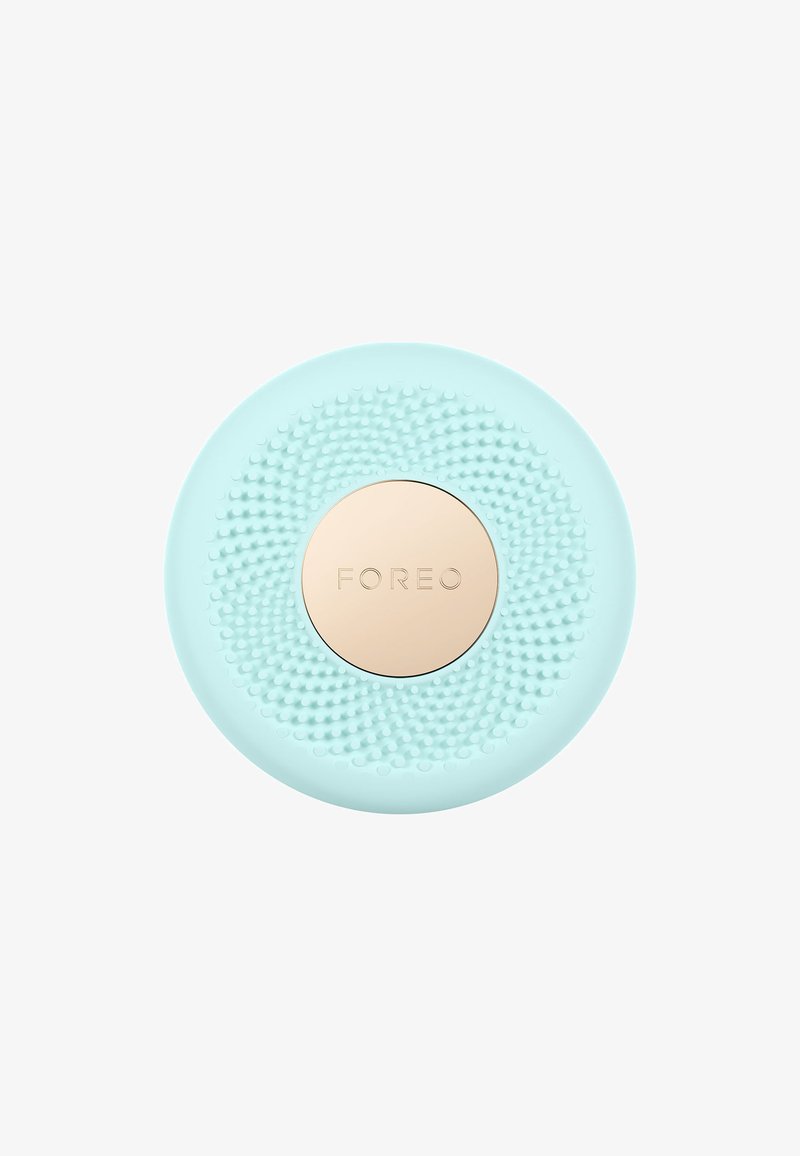 Silicone facial cleansing device in mint green with textured surface. Gold circular center features brand logo. Round shape, compact design.