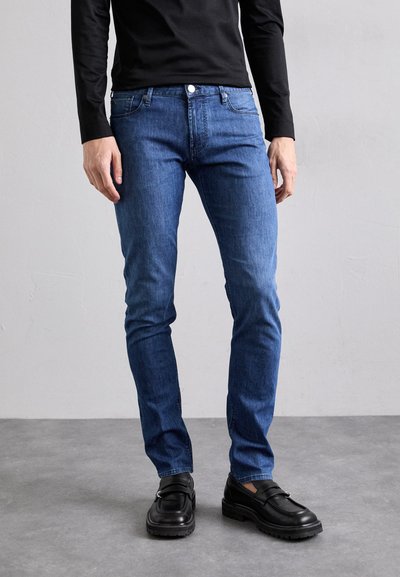 Schmale blaue Denim-Jeans mit glatter Textur, ausgestattet mit einem Knopfverschluss, fünf Taschen und subtilen Steppnähte. Dazu werden schwarze Slipper getragen.