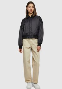 Jachetă bomber din satin negru cu guler din blană, având fermoar frontal și manșete elastice, combinată cu pantaloni largi crema și adidași albi.