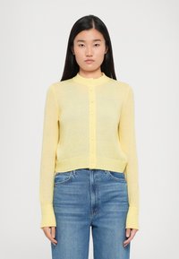 Gul, lett strikket cardigan med rund krage, knappelukking foran og lange ermer. Bæreres over blå denimjeans.