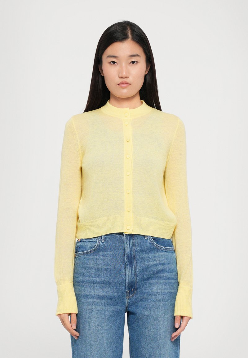 Gul, lett strikket cardigan med rund krage, knappelukking foran og lange ermer. Bæreres over blå denimjeans.