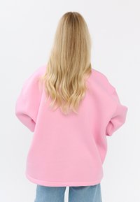 Felpa rosa oversize in morbido tessuto, caratterizzata da una vestibilità rilassata, spalle scese e scollatura a girocollo, abbinata a jeans blu chiaro.