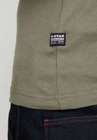 Oliivinvihreä t-paita, joka on valmistettu puuvillasta ja jossa on slim fit -malli, ja helman kohdalla on musta tägi. Tummat farkut, joissa on oranssit tikkaukset, näkyvät osittain.