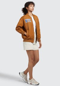 Giacca bomber marrone con "NYC" in blu, t-shirt bianca, pantaloni corti crema e sneakers beige con dettagli azzurri. Sfondo semplice.