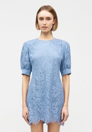 Γυναίκα που φοράει ένα ανοιχτό μπλε φόρεμα από δαντέλα με φουσκωτά κοντά μανίκια και κυματιστό τελείωμα, στέκεται μπροστά σε ένα απλό φωτεινό φόντο.