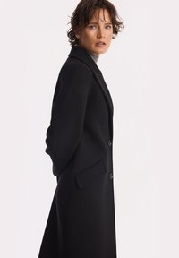 Kobieta z krótkimi, kręconymi włosami, ubrana w długi czarny płaszcz i szary sweter z golfem, stojąca bokiem na jednolitym jasnym tle.