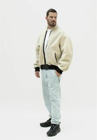 Beige faux fur bomberjakke med en sort ribbet kant og manchetter, parret med lyseblå jeans og sorte sneakers. Lukkes med lynlås foran.