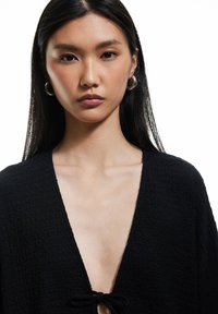 Top nero testurizzato con scollatura a V profonda, caratterizzato da un dettaglio con nodo frontale. La modella ha capelli lunghi e indossa grandi orecchini a cerchio in argento.