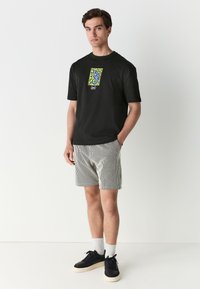 Black cotton t-shirt with graphic print, paired with striped black and white shorts, and black sneakers with white soles.