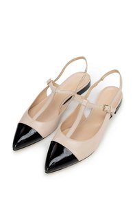 Zapatos de piel beige tipo slingback con puntera afilada, que presentan una puntera negra brillante, diseño de tira en T y detalle de hebilla en el tobillo. Suela plana.