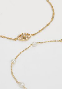 Gold necklace with twisted chain and an eye-shaped pendant adorned with pearls and small stones; second chain features pearl accents.