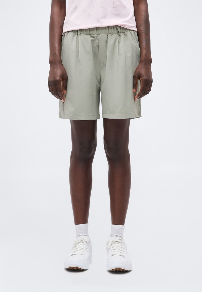 Άτομο που φοράει σορτς με ελαστική μέση σε ανοιχτό πράσινο χρώμα, λευκές κάλτσες, λευκά αθλητικά παπούτσια και ανοιχτό ροζ μπλουζάκι, στέκεται μπροστά σε απλό ανοιχτό φόντο.