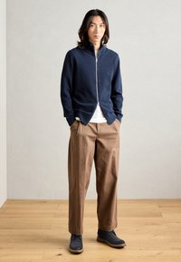 Maglione navy con zip, camicia beige a girocollo sotto, pantaloni beige a gamba larga e scarpe navy. Tessuto liscio, vestibilità casual, design minimalista.