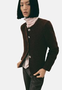 Cardigan en laine bordeaux avec une surface texturée, trois boutons argentés, ouvert à l'avant et coupe ajustée. Superposé à un col roulé rose pâle.