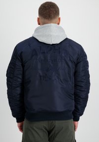 Marinfärgad bomberjacka med kviltad textur och ribbad mudd. Har broderad logotyp och en grå huva som tittar fram under.