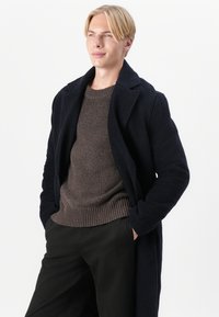 Merinvärldö som har en texturerad tyg, ficklappar och en avslappnad passform, lager på en brun stickad tröja och svarta byxor.