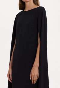 Musta mekko, jossa on venepääntie, viittamaiset hihat ja sileä, nestemäinen tekstuuri. Minimalistinen muotoilu ilman kuvioita tai koristeluja.
