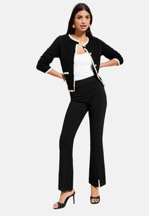 Frau trägt eine schwarze, strukturierte Strickjacke mit weißem Besatz, ein weißes Tanktop, schwarze, hoch taillierte Hosen mit vorderen Schlitzen und schwarze Sandalen mit hohem Absatz.