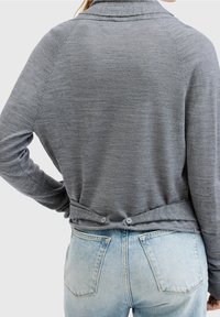 Szary sweter z długim rękawem o strukturze. Posiada kołnierz szalowy oraz dwa guziki na dolnej części pleców, w zestawie z jasnoniebieskimi dżinsami.