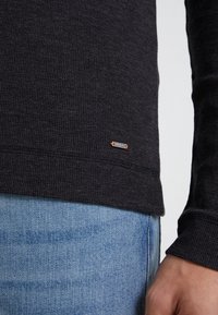 Tummanharmaa pitkähihainen paita, jossa on teksturoitu neulos ja pieni merkkilappu helmassa, yhdistettynä siniseen denimmateriaalin alaosaan.