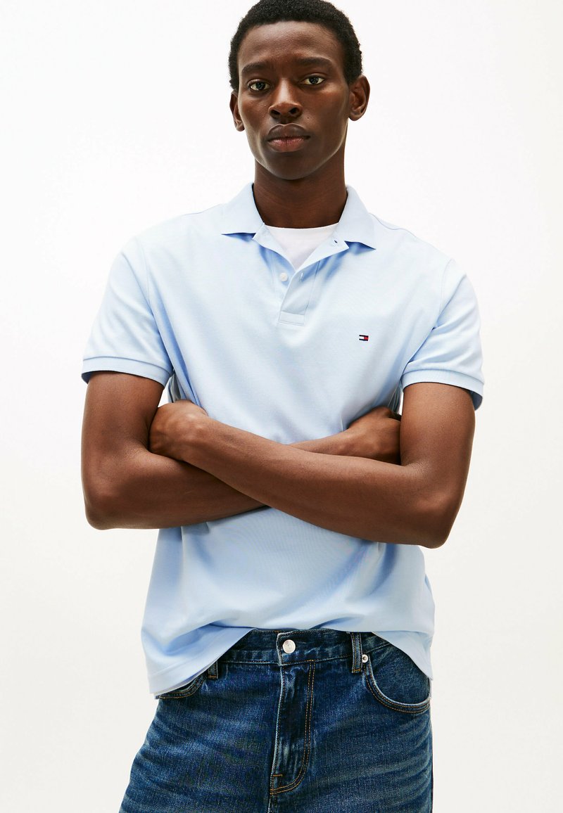 Lichtblauw poloshirt met korte mouwen en een kraag, met een klein logo op de borst. Draag het over een donkere jeans met de armen gekruist.