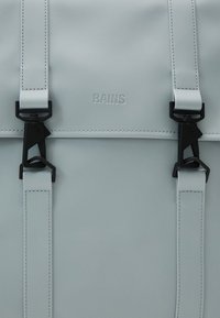 Sac à dos imperméable gris clair avec deux fermoirs noirs et des sangles verticales, embossé du nom de la marque "RAINS" au centre.